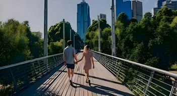 A couple walking hand in hand across a bridge with trees on either side and a cityscape ahead.