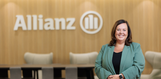 Julie Mitchell stands in the foreground wearing a green suit jacket. In the background are office tables and chairs against a wall that features the Allianz logo in white.