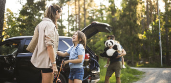 Mother and daughter standing beside an electric vehicle being charged whilst father unloads second child and large panda soft toy from vehicle