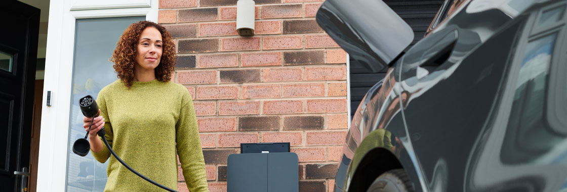 A woman at a residential location approaching an electric vehicle with a charging cable in hand, ready to plug it in.