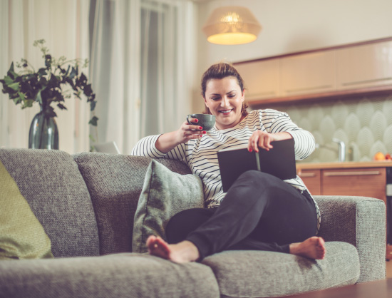 A smiling woman seated in a living room setting reading a book and holding a hot beverage in one hand