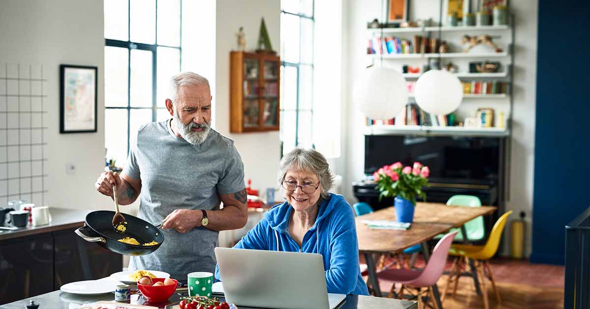 Senior couple in a kitchen, woman seated looking at a laptop, man standing with a frypan.