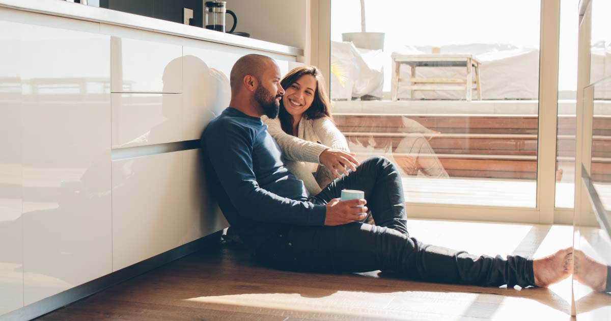 Heterosexual middle-aged couple seated leaning against a wall in casual conversation holding coffee mugs