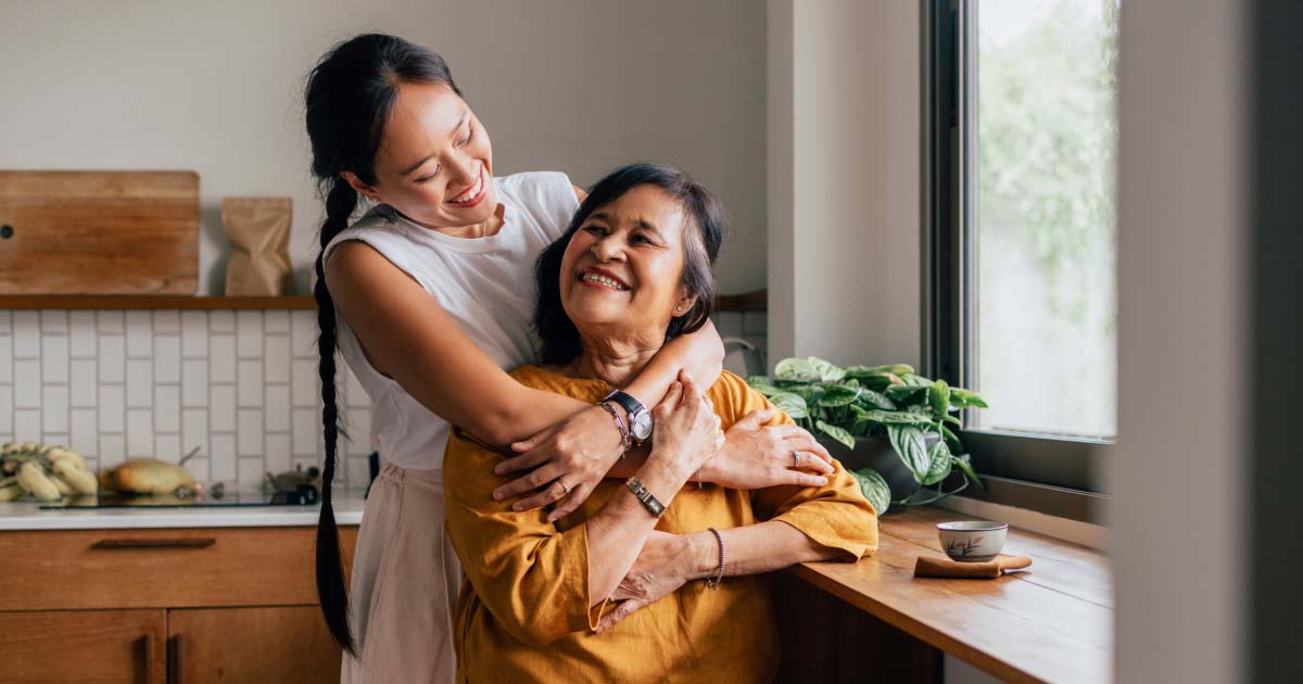 A woman hugs an older woman from behind in a kitchen with wooden cabinets and a potted plant on the window sill.