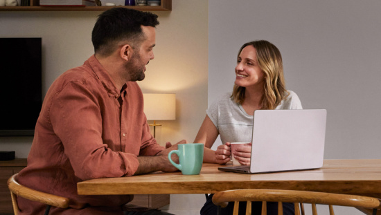 Male and female in discussion at a dining table seated next to an open laptop holding coffee mugs